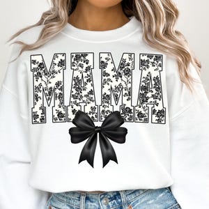 May include: White sweatshirt with the word "MAMA" in large, block letters filled with a black floral pattern. A large black bow is centered below the text. The sweatshirt is a casual, everyday style.