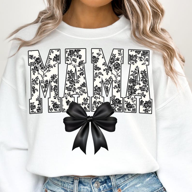 May include: White sweatshirt with the word "MAMA" in large, block letters filled with a black floral pattern. A large black bow is centered below the text. The sweatshirt is a casual, everyday style.