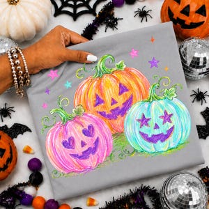 May include: A gray t-shirt featuring a colorful graphic of three jack-o'-lanterns in orange, pink, and light blue. The pumpkins have purple carved faces with heart and star-shaped eyes, surrounded by small pink, purple, and blue stars.