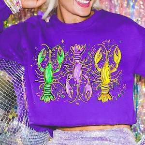 May include: A purple sweatshirt featuring a graphic of three colorful lobsters. The lobsters are green, purple, and yellow, with gold accents and a speckled background. The sweatshirt is a solid purple color.