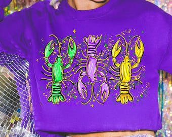 May include: A purple sweatshirt featuring a graphic of three colorful lobsters. The lobsters are green, purple, and yellow, with gold accents and a speckled background. The sweatshirt is a solid purple color.