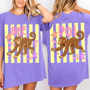 May include: A purple t-shirt with a cartoon monkey design. The design includes the words "SEE" and "DO!" in pink, along with stars and other shapes. The background has yellow and white stripes.