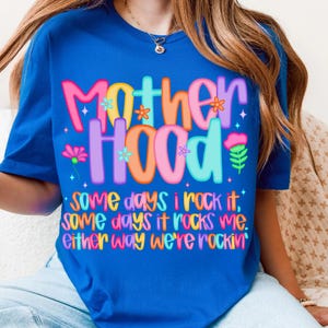 May include: A royal blue t-shirt with the word "MOTHERHOOD" in colorful, bubble letters. The shirt also features the text "Some days I rock it, some days it rocks me. Either way we're rockin'" in a playful font.