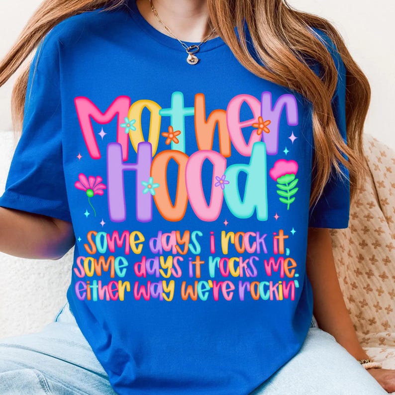 May include: A royal blue t-shirt with the word "MOTHERHOOD" in colorful, bubble letters. The shirt also features the text "Some days I rock it, some days it rocks me. Either way we're rockin'" in a playful font.