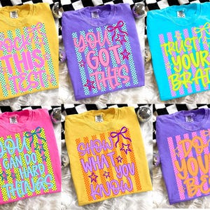 Pode incluir: Seis camisetas coloridas com frases motivacionais. As camisetas são amarelas, roxas, rosas e azuis. Cada camiseta tem uma frase diferente em uma fonte ousada e colorida, como "Rock This Test" e "Confie no seu cérebro".