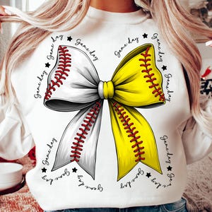 May include: White sweatshirt featuring a large bow design. The bow is split into two halves, one white and the other yellow, each resembling a baseball and softball. Red stitching details the bow, with the words "Game Day" and stars around the bow.