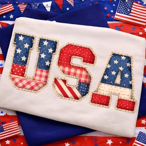 May include: White t-shirt with the word "USA" appliqued in red, white, and blue fabric. The letters are filled with star and stripe patterns, and the edges are outlined with a tan stitch. The shirt is surrounded by patriotic decorations.