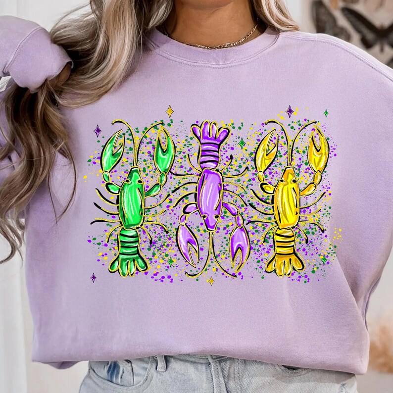 May include: A light purple sweatshirt featuring a colorful graphic of three crawfish. The crawfish are green, purple, and yellow with gold accents, and are surrounded by colorful confetti.