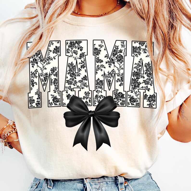 May include: Cream-colored t-shirt with the word "MAMA" in large, black floral print letters. A black bow is centered below the text. The shirt is a casual style.
