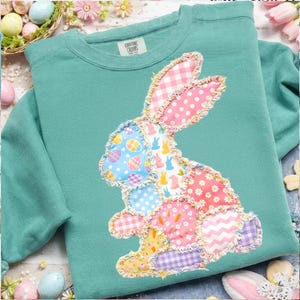 May include: A teal sweatshirt featuring a patchwork bunny design. The bunny is composed of various patterned fabrics, including polka dots, gingham, and floral prints. The sweatshirt has a crew neck and long sleeves, perfect for Easter.