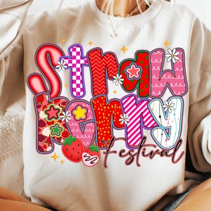 May include: Cream-coloured sweatshirt with a colourful graphic that reads "Strawberry Festival." The text features various patterns, including strawberries, stars, and floral accents. The design is outlined in black.