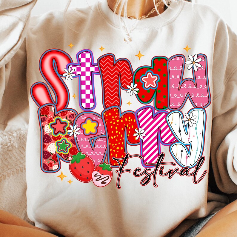 May include: Cream-coloured sweatshirt with a colourful graphic that reads "Strawberry Festival." The text features various patterns, including strawberries, stars, and floral accents. The design is outlined in black.