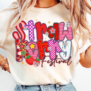 May include: A cream-coloured t-shirt with the words "Strawberry Festival" in a colourful, whimsical design. The text features strawberries, flowers, and various patterns in red, pink, and purple. The shirt is a casual, everyday wear item.