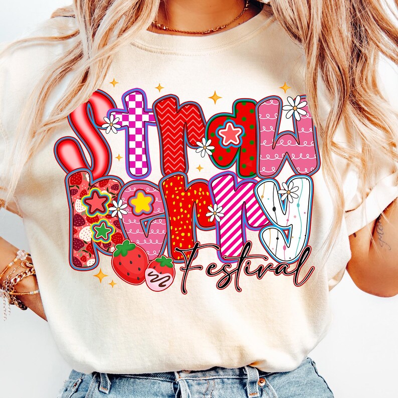 May include: A cream-coloured t-shirt with the words "Strawberry Festival" in a colourful, whimsical design. The text features strawberries, flowers, and various patterns in red, pink, and purple. The shirt is a casual, everyday wear item.
