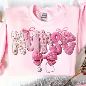 May include: A light pink sweatshirt with the word "NURSE" in a decorative, textured design. The letters are embellished with bows, flowers, and patterns. A large pink bow and a stethoscope are below the word.