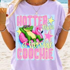 May include: A light purple t-shirt featuring a graphic of a green alligator wearing pink sunglasses while lounging in a pink flamingo pool float. The design includes yellow stars and the text "HOTTER than a hoochie COOCHIE" in pink and yellow.