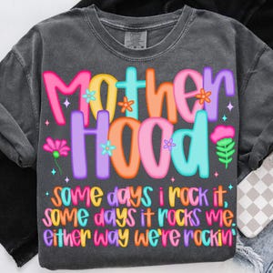 May include: Dark gray t-shirt with the word "Motherhood" in colorful bubble letters. The shirt has a floral design and the text "Some days I rock it, some days it rocks me. Either way we're rockin'".