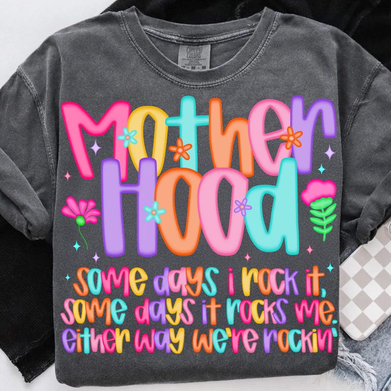 May include: Dark gray t-shirt with the word "Motherhood" in colorful bubble letters. The shirt has a floral design and the text "Some days I rock it, some days it rocks me. Either way we're rockin'".