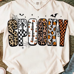 May include: Cream-colored t-shirt with the word "SPOOKY" in a variety of patterns, including leopard print, checkerboard, and pumpkins. The design includes bats, spiderwebs, and small spiders, perfect for Halloween.