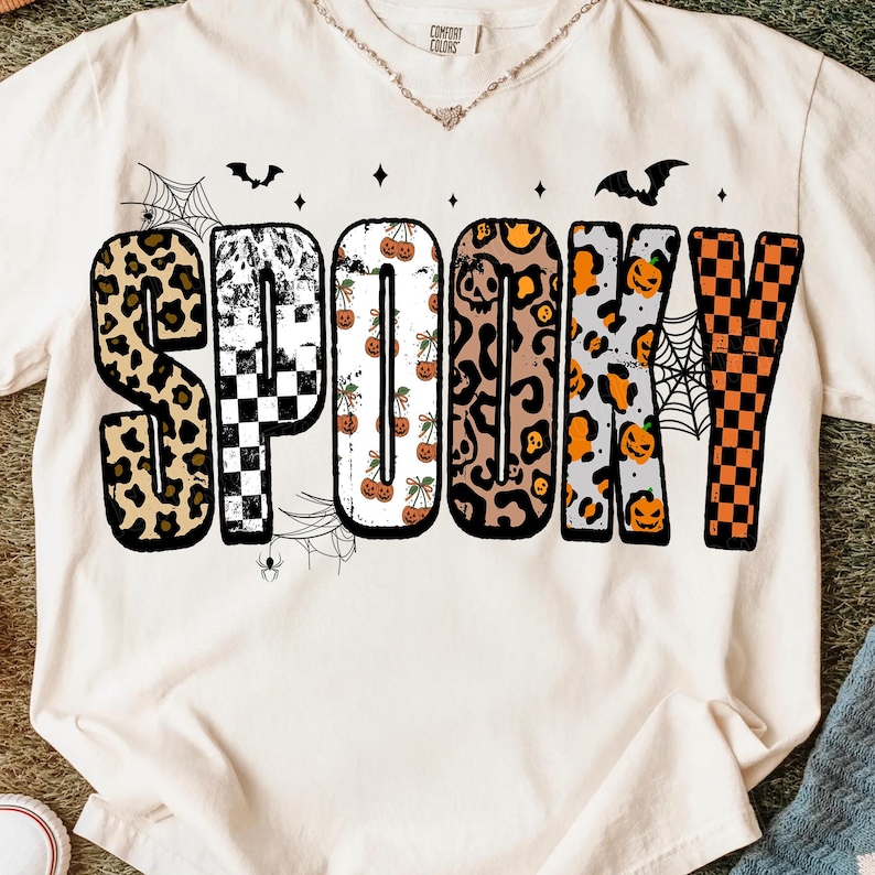 May include: Cream-colored t-shirt with the word "SPOOKY" in a variety of patterns, including leopard print, checkerboard, and pumpkins. The design includes bats, spiderwebs, and small spiders, perfect for Halloween.