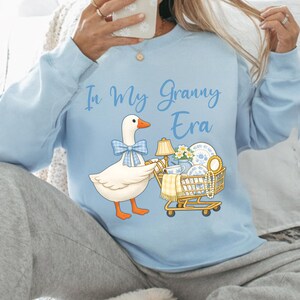 Puede incluir: Sudadera azul claro con la frase "In My Granny Era". El gr&aacute;fico presenta un ganso de dibujos animados con una pajarita azul, empujando un carrito de compras lleno de art&iacute;culos vintage, incluyendo una l&aacute;mpara y vajilla.