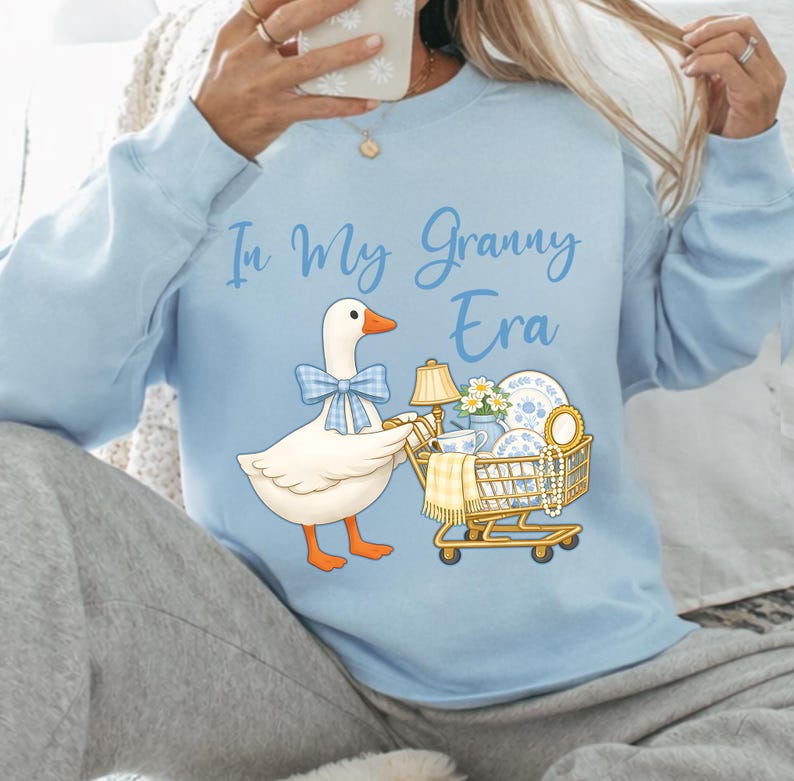 Puede incluir: Sudadera azul claro con la frase "In My Granny Era". El gr&aacute;fico presenta un ganso de dibujos animados con una pajarita azul, empujando un carrito de compras lleno de art&iacute;culos vintage, incluyendo una l&aacute;mpara y vajilla.