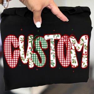 Pu&ograve; includere: Felpa nera con la scritta "CUSTOM" in grandi lettere festive. Le lettere sono riempite con motivi a quadri rossi e verdi, disegni floreali e accentate con fiocchi e bacche, perfetta per le festivit&agrave;.