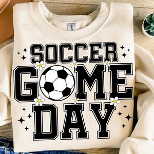 May include: Cream-colored sweatshirt with the words "SOCCER GAME DAY" in black block letters. A soccer ball graphic is in the center, with small white flowers and black star accents. The sweatshirt is a casual, sporty style.