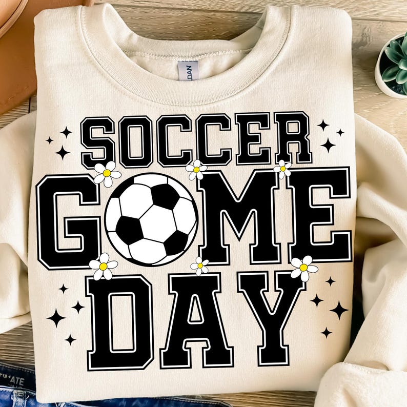 May include: Cream-colored sweatshirt with the words "SOCCER GAME DAY" in black block letters. A soccer ball graphic is in the center, with small white flowers and black star accents. The sweatshirt is a casual, sporty style.