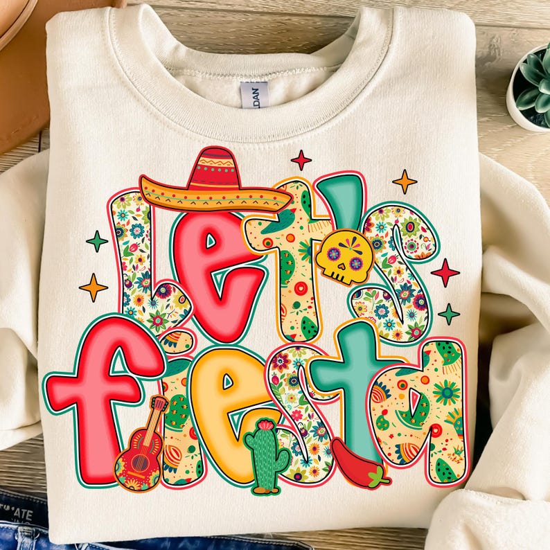 May include: Cream-colored sweatshirt with the words "Let's Fiesta" in a colorful, festive design. The design includes a sombrero, guitar, cactus, chili pepper, and a skull, all with floral patterns. The sweatshirt is a casual, comfortable garment.