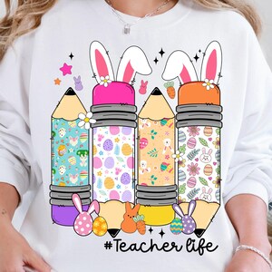 Op de afbeelding: Wit sweatshirt met een afbeelding van vier potloden versierd met paasmotieven en konijnenoren. De tekst "#Teacher life" is onder de potloden gedrukt.
