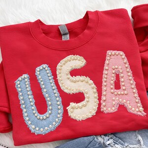 K&ouml;nnte beinhalten: Ein rotes Sweatshirt mit dem Wort "USA" auf der Vorderseite gestickt. Die Buchstaben sind blau, cremefarben und rosa, jeweils mit kleinen, wei&szlig;en Perlen umrandet. Das Sweatshirt hat einen Rundhalsausschnitt und lange &Auml;rmel.
