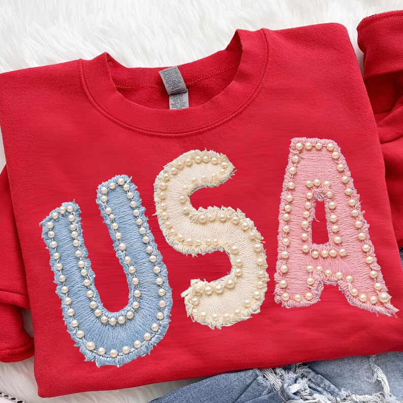 K&ouml;nnte beinhalten: Ein rotes Sweatshirt mit dem Wort "USA" auf der Vorderseite gestickt. Die Buchstaben sind blau, cremefarben und rosa, jeweils mit kleinen, wei&szlig;en Perlen umrandet. Das Sweatshirt hat einen Rundhalsausschnitt und lange &Auml;rmel.
