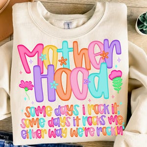 May include: Cream-colored sweatshirt with the words "Motherhood" in colorful, bubble letters. The text is decorated with flowers and stars. Below, the sweatshirt reads "Some days I rock it, some days it rocks me, either way we're rockin."