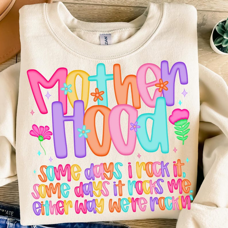 May include: Cream-colored sweatshirt with the words "Motherhood" in colorful, bubble letters. The text is decorated with flowers and stars. Below, the sweatshirt reads "Some days I rock it, some days it rocks me, either way we're rockin."