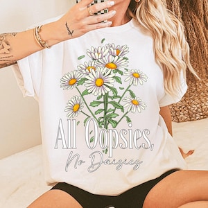 K&ouml;nnte beinhalten: Wei&szlig;es T-Shirt mit einer G&auml;nsebl&uuml;mchen-Bouquet-Grafik mit gelben Zentren und wei&szlig;en Bl&uuml;tenbl&auml;ttern. Der Text "All Oopsies, No Daisies" ist unter den Blumen in einer Schreibschrift gedruckt. Das Shirt hat kurze &Auml;rmel.