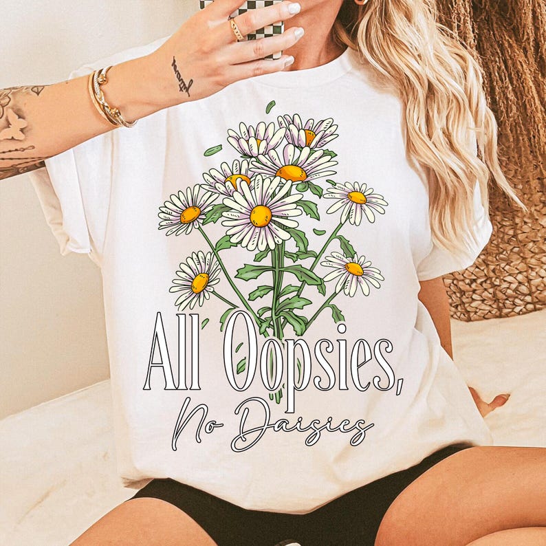 K&ouml;nnte beinhalten: Wei&szlig;es T-Shirt mit einer G&auml;nsebl&uuml;mchen-Bouquet-Grafik mit gelben Zentren und wei&szlig;en Bl&uuml;tenbl&auml;ttern. Der Text "All Oopsies, No Daisies" ist unter den Blumen in einer Schreibschrift gedruckt. Das Shirt hat kurze &Auml;rmel.