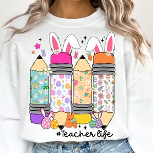 Op de afbeelding: Wit sweatshirt met een afbeelding van drie kleurrijke potloden met konijnenoren en paasei-patronen. De tekst "#Teacher life" is onder de potloden gedrukt.
