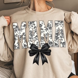 May include: Beige sweatshirt with the word "MAMA" in large, bold letters. The letters are filled with a black and white floral pattern. A large black bow is centered below the word "MAMA".