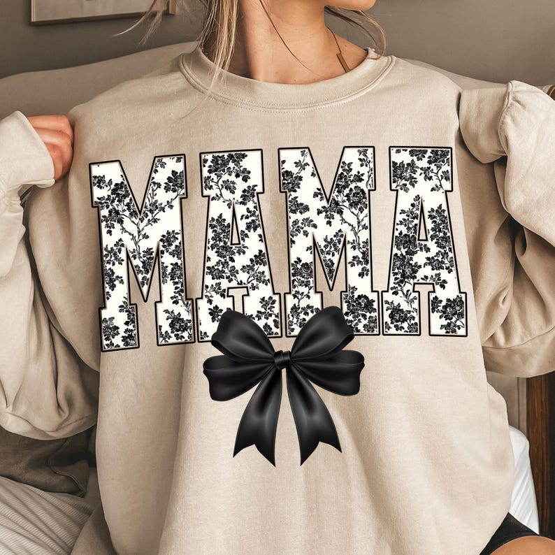 May include: Beige sweatshirt with the word "MAMA" in large, bold letters. The letters are filled with a black and white floral pattern. A large black bow is centered below the word "MAMA".