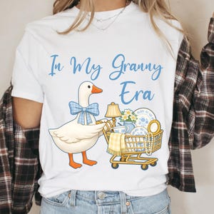 Puede incluir: Camiseta blanca con el texto "In My Granny Era" en escritura azul. Un ganso de dibujos animados con una pajarita azul empuja un carrito de compras lleno de objetos vintage, incluyendo una l&aacute;mpara, vajilla y flores.