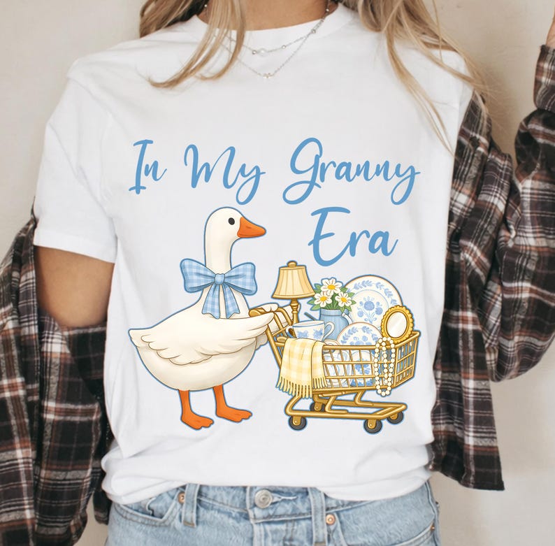 Puede incluir: Camiseta blanca con el texto "In My Granny Era" en escritura azul. Un ganso de dibujos animados con una pajarita azul empuja un carrito de compras lleno de objetos vintage, incluyendo una l&aacute;mpara, vajilla y flores.