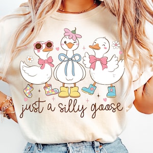 May include: A cream-coloured t-shirt with a cartoon design of three geese. Each goose has a bow, boots, and different accessories, including sunglasses and flowers. The text "just a silly goose" is written in a cursive font below the geese.