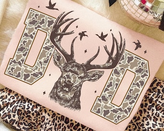 May include: A light pink sweatshirt with the word "DAD" in a camouflage pattern. A detailed illustration of a deer with large antlers is centered between the letters. Small birds and stars are scattered around the design.