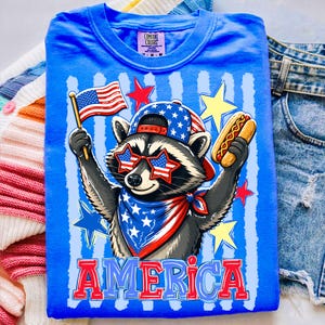 Könnte beinhalten: Ein leuchtend blaues T-Shirt mit einer Grafik eines Waschbären, der eine sternenbesetzte Baseballkappe, sternförmige Sonnenbrillen und ein Halstuch trägt. Der Waschbär hält eine US-Flagge und einen Hotdog. Das Wort AMERICA steht in Rot und Blau.