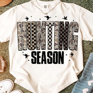 Puede incluir: Camiseta color crema con las palabras "HUNTING SEASON" en un dise&ntilde;o de camuflaje y cuadros negros. La camiseta presenta peque&ntilde;os gr&aacute;ficos de p&aacute;jaros y estrellas. Un collar es visible en el escote.
