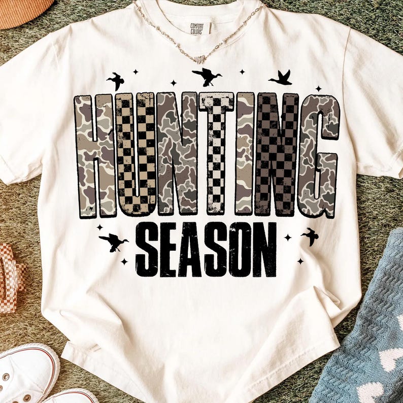 Puede incluir: Camiseta color crema con las palabras "HUNTING SEASON" en un dise&ntilde;o de camuflaje y cuadros negros. La camiseta presenta peque&ntilde;os gr&aacute;ficos de p&aacute;jaros y estrellas. Un collar es visible en el escote.