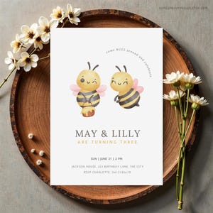 May include: A white invitation card featuring illustrations of two cartoon bees, honey, and text. The invitation reads "MAY & LILLY ARE TURNING THREE" with event details. The card is on a wooden tray with white flowers.