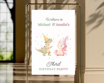 Op de afbeelding: Een gouden metalen frame houdt een wit bord met de tekst "Welcome to Michael & Amalia's Third BIRTHDAY PARTY!". Het bord toont illustraties van een dinosaurus en een eenhoorn met feesthoeden. De achtergrond is lichtbeige.