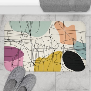 May include: Rectangular bath mat with an abstract design. The mat features a cream background with colorful shapes in teal, peach, yellow, black, and purple, overlaid with black line art. Gray slippers are at the bottom.